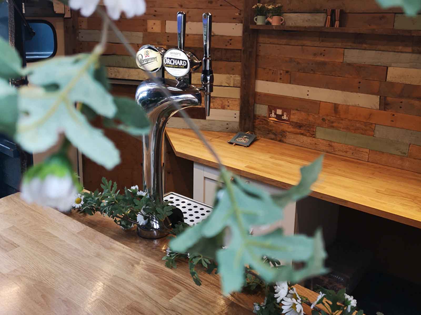 A close up view inside The Brew Box mobile bar, showing a beer pump, wooden work surfaces and floral decorations